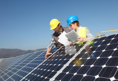 Ein Mann mit gelbem Helm zeigt einer jungen Frau mit blauem Helm etwas auf einem Solarpanel vor blauem Himmel.
