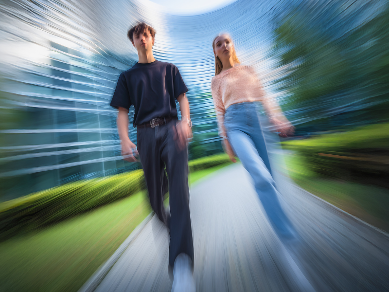 Due giovani adulti camminano verso la telecamera in un moderno parco, con uno svettante edificio di vetro sullo sfondo. L'immagine è caratterizzata da una forte sfocatura dovuta al movimento e da un effetto vorticoso.