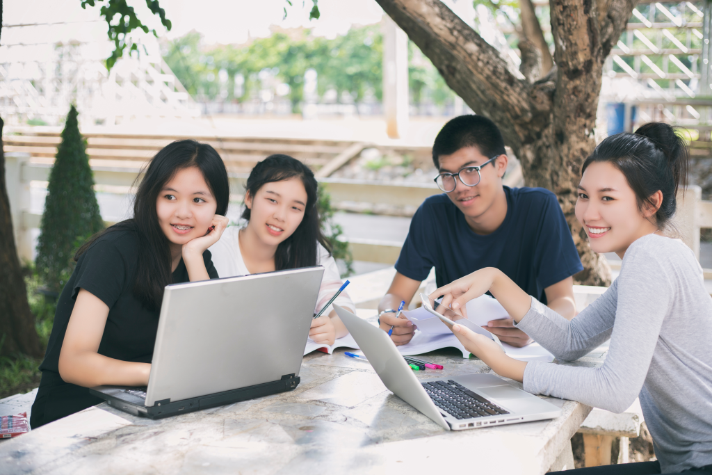 Gruppo di studenti asiatici che utilizzano tablet e notebook e condividono le idee per lavorare sul prato del campus